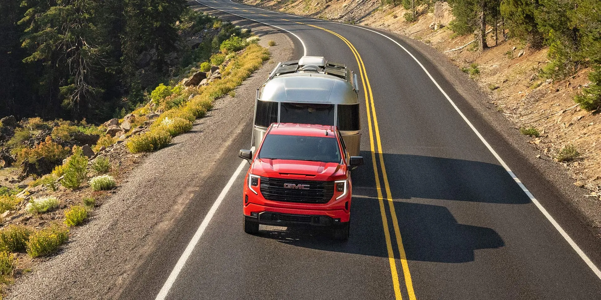 GMC Sierra 1500 2026 rouge remorquant une roulotte sur une route de montagne.