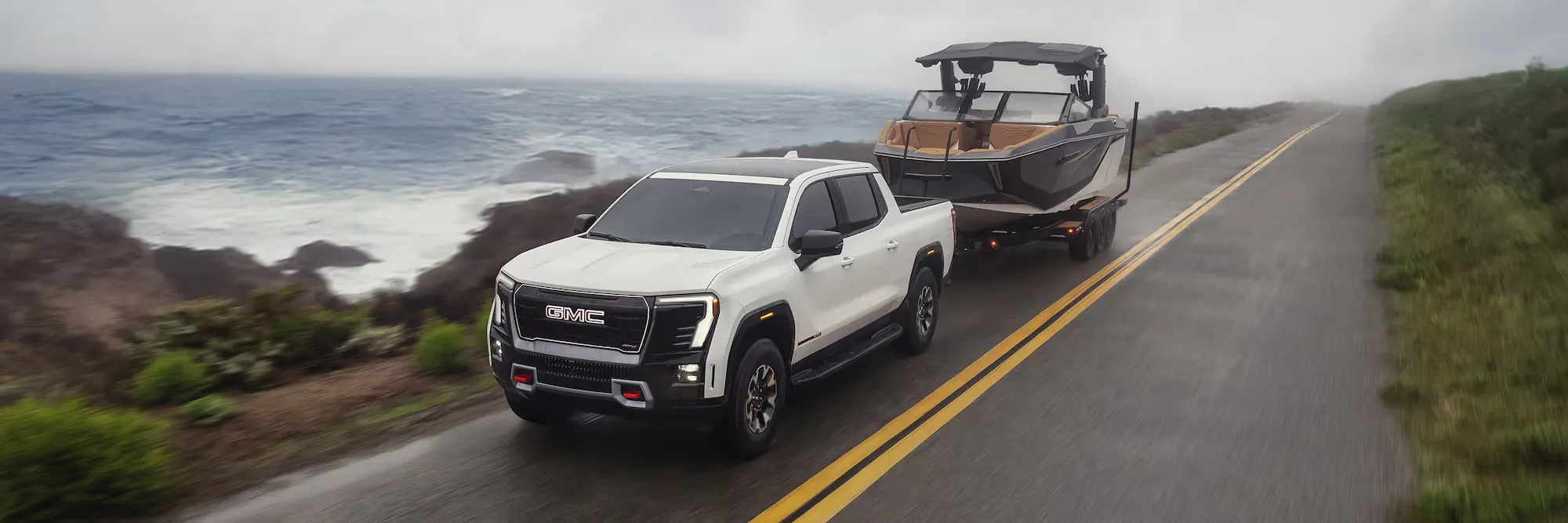 GMC Sierra EV 2026 blanc remorquant un bateau sur une route côtière.